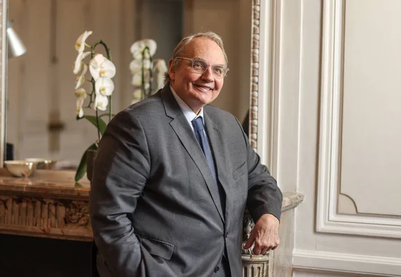Jean-Louis Bourlanges souriant dans un intérieur élégant avec des orchidées en arrière-plan.