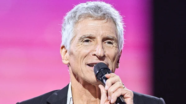 Nagui micro en main, portant une veste sur chemise claire, devant un fond rose.