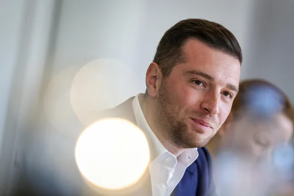 Portrait de Jordan Bardella portant une veste sombre et une chemise blanche, regardant la caméra avec un léger sourire.