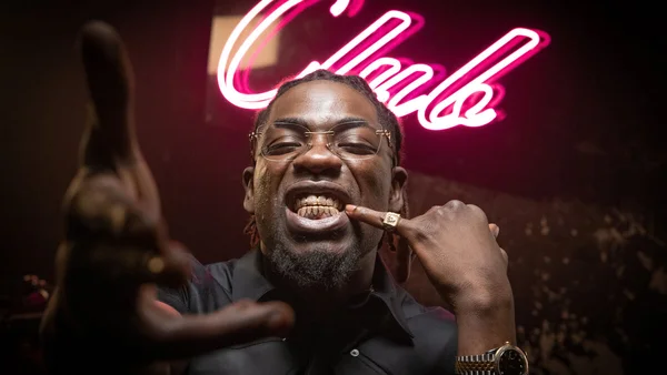 Portrait du rappeur Gazo avec des grills et des lunettes devant un fond néon rose.