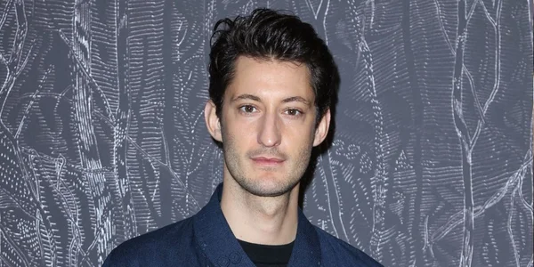 Pierre Niney en veste bleue foncée devant un fond décoratif gris.