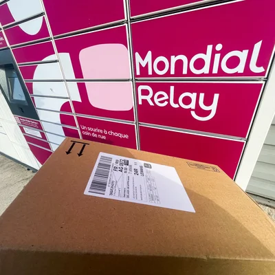 Un paquet carton déposé devant un locker Mondial Relay affichant la devise 'Un sourire à chaque coin de rue'.