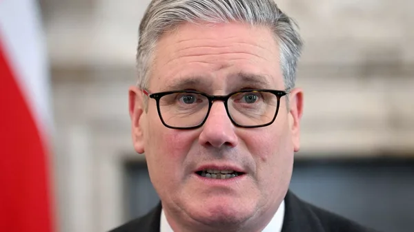 Keir Starmer, en costume sombre et lunettes noires, portrait rapproché devant un drapeau rouge et blanc.