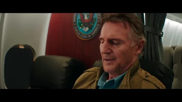 Un homme assis dans un avion gouvernemental américain, portant une veste beige et une chemise turquoise.