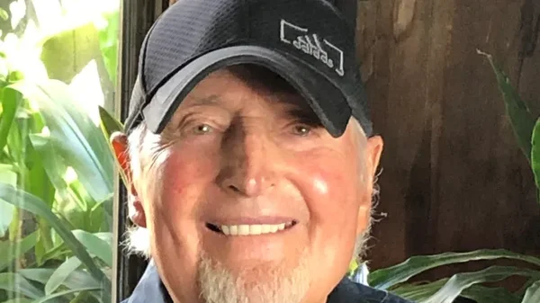 Un homme souriant avec une casquette noire et une chemise bleue devant des plantes.