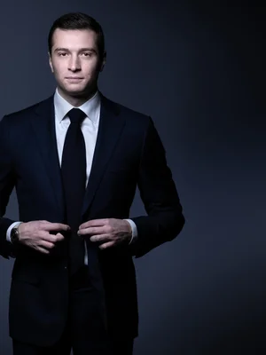 Jordan Bardella en costume debout sur fond gris.