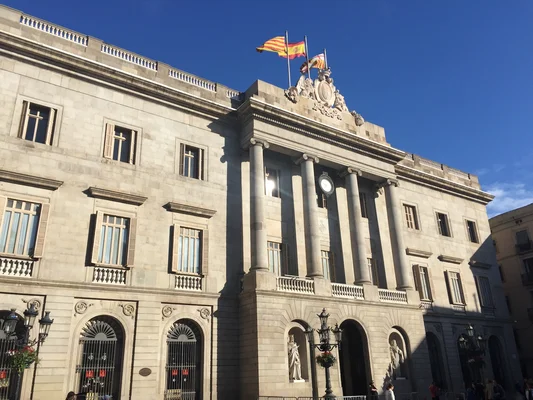 La Mairie de Barcelone, bâtiment historique en pierre avec colonnes et arcs, surplombant une place publique fréquentée par des passants.
