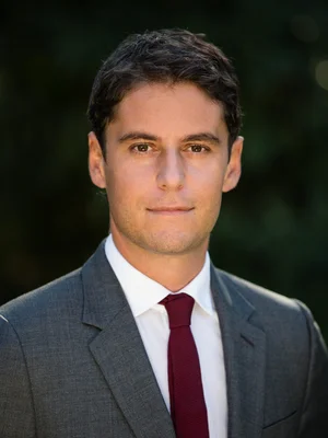 Gabriel Attal posant en costume gris et cravate bordeaux.