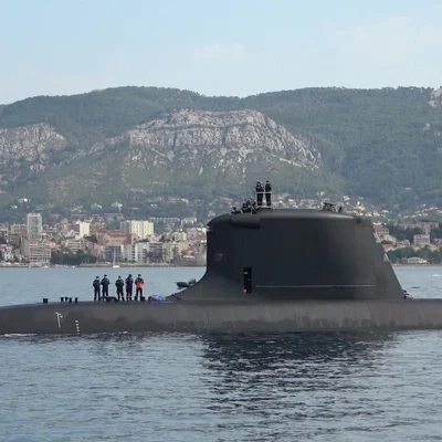 Le sous-marin Le Suffren avec des membres d'équipage, vu devant un paysage urbain et montagneux.