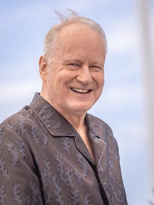 Stellan Skarsgård souriant en chemise brune à motifs, devant un ciel bleu.