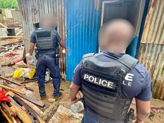 Des policiers face à une habitation en tôle ondulée dans une zone difficile d'accès.