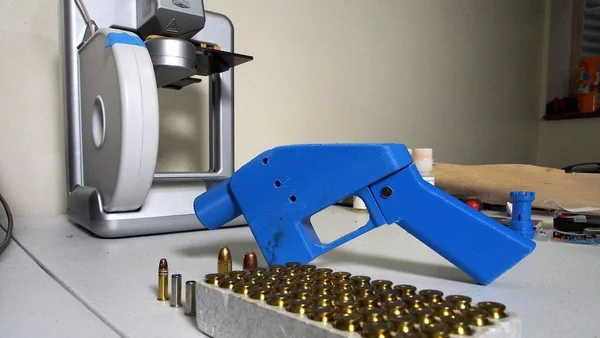 Un pistolet bleu imprimé en 3D avec des douilles en laiton à côté d'une imprimante 3D.