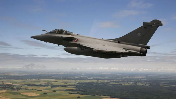 Un chasseur Dassault Rafale en vol au-dessus d'une campagne boisée sous un ciel nuageux.