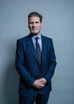 Portrait officiel de Keir Starmer en costume bleu, les mains jointes devant un fond gris
