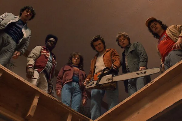 Un groupe de six personnes se tient sur une structure en bois, dont une arme une tronçonneuse, dans une ambiance années 80.