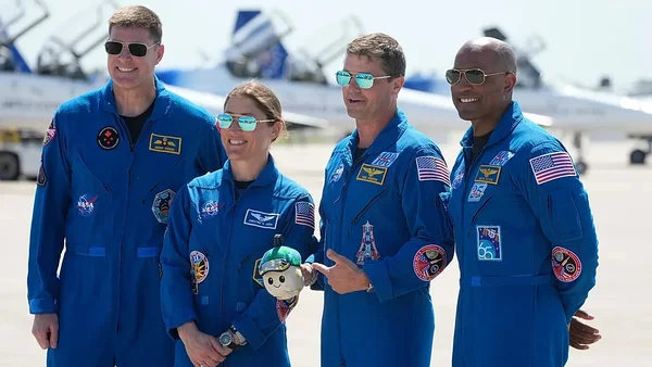 L'équipage Artemis II en bleus de vol NASA sur un aérodrome avec des avions en arrière-plan, dont un membre tenant une peluche.