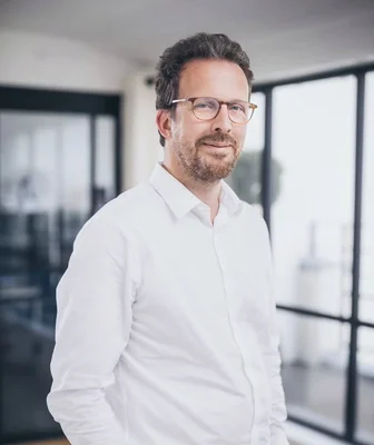 François Durvye en chemise blanche, barbu et portant des lunettes, dans un intérieur lumineux.