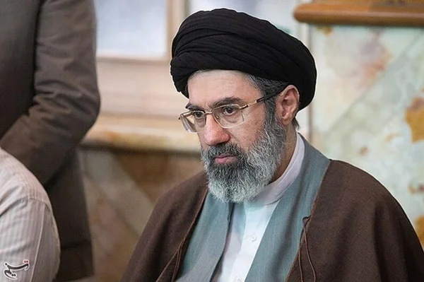 Portrait de Mojtaba Khamenei assis, identifiable à sa barbe grise, son turban noir et ses lunettes.