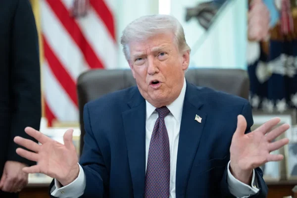 Donald Trump assis dans un bureau officiel, gestiquant avec les mains devant un drapeau américain.