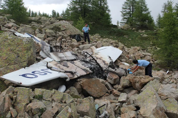 Les restes d'un avion accidenté dans un décor rocheux, examinés par deux personnes.