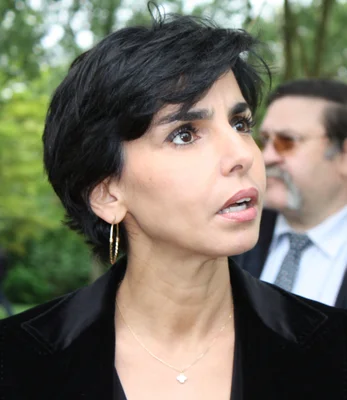 Rachida Dati vêtue d'un blazer noir et de boucles d'or, regardant vers le haut lors d'un événement en extérieur.