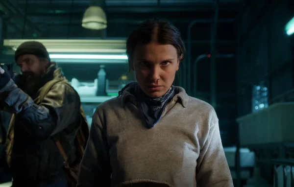 Une femme en beige et un homme barbu dans un décor industriel sombre.