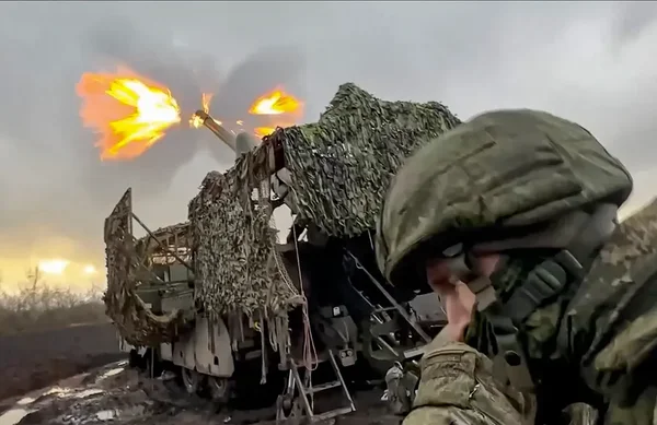 Un soldat regardant un véhicule militaire tirer lors d'un conflit, fumée et éclats en arrière-plan.