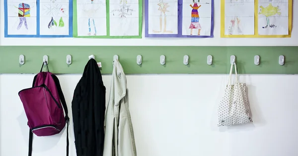 Un porte-manteau d'école avec cartables et vestes, sous des dessins d'enfants encadrés.