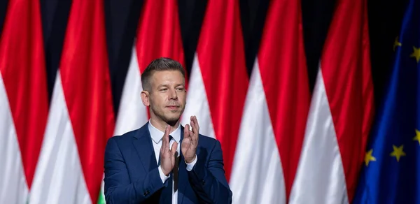Un homme en costume applaudissant devant une scène décorée de drapeaux nationaux et européens.
