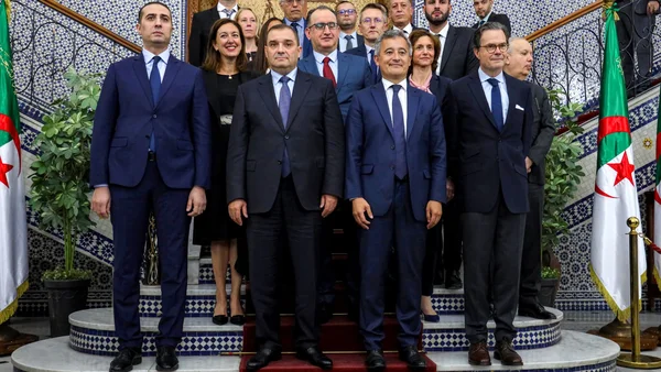 Portrait de Gérald Darmanin, en costume bleu, devant un mur ornemental.