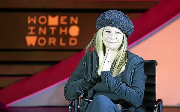 Barbra Streisand, bonnet gris et veste noire, mains jointes, devant un mur 'WOMEN IN THE WORLD'.