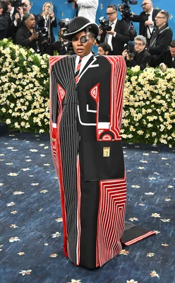 Une tenue avant-gardiste rayée noir et rouge sur le tapis rouge du Met Gala 2025, devant un mur de fleurs blanches.