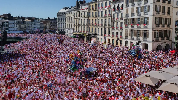 Foule dense lors des Fêtes de Bayonne, place entourée d'immeubles anciens et de chars décorés.