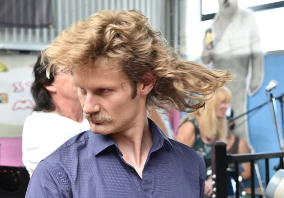 Un homme à la coupe mulet blonde et à la moustache, dans une pose naturelle.