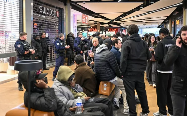 Scène intérieure dans un centre commercial : des policiers en uniforme se tiennent près d'une boutique Swatch tandis que des clients sont assis ou debout à proximité, après le lancement chaotique.