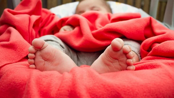 Gros plan sur les pieds nus d’un bébé endormi sous une couverture rouge.