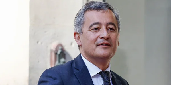 Gérald Darmanin saluant devant des drapeaux, dont le drapeau français, lors de son retour d'Alger.