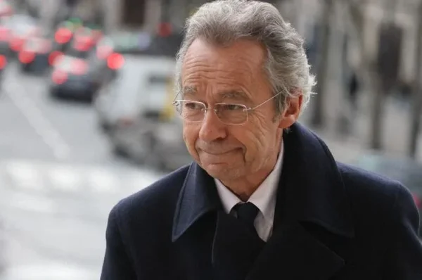 Michel Denisot, cheveux gris et lunettes, en manteau sombre et cravate, debout dans une rue.