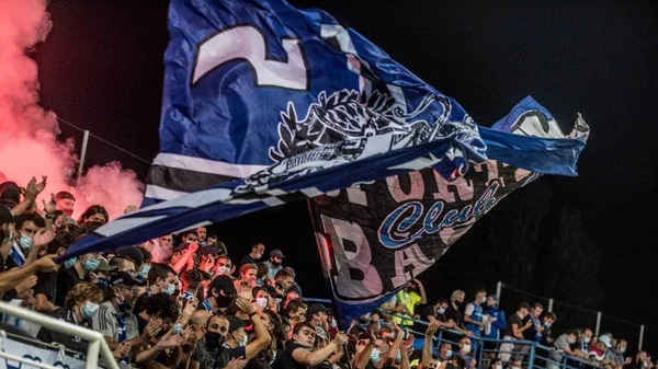 Drapeau géant du SC Bastia brandi par les supporters, avec des fumigènes rouges en arrière-plan.