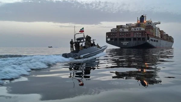 Un bateau rapide battant pavillon iranien dans le détroit d'Ormuz, avec un porte-conteneurs en arrière-plan.