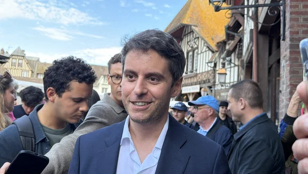 Gabriel Attal, souriant, entouré de la foule et de la presse dans une rue ensoleillée aux maisons à colombages.