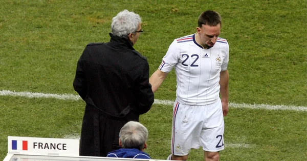 Raymond Domenech et Franck Ribéry se serrant la main sur le terrain, avec un drapeau français en arrière-plan.