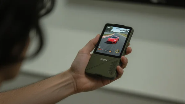 Vue rapprochée de la main d'un joueur tenant la ROG Ally 2 en mode portable, écran affichant un jeu de course avec une voiture rouge, lumière tamisée d'une chambre