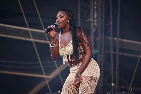 Aya Nakamura en concert, vêtue d'une tenue crochet beige à franges et bijoux dorés.
