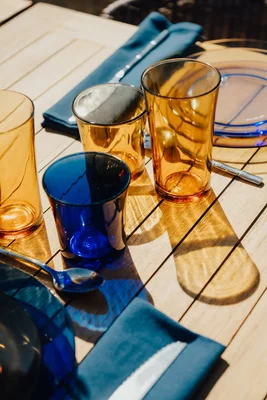Collection de gobelets et assiettes Duralex, aux teintes ambre et bleu cobalt, sur une table en bois éclairée par le soleil.