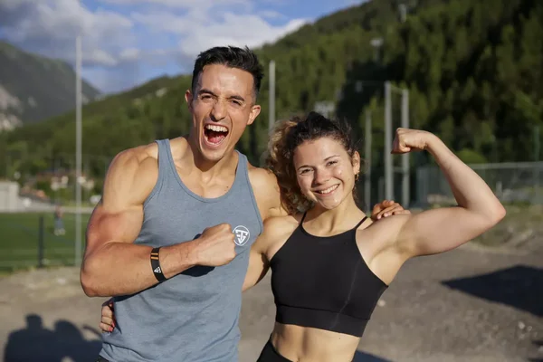Tibo InShape et Juju Fitcats affichant leur musculature devant un paysage montagneux.