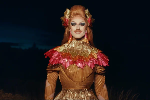 Pattie Gonia en robe dorée à col de plumes, dans un décor crépusculaire