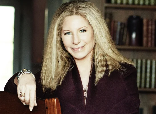Barbra Streisand lors d'une séance photo, veste violette et bibliothèque en arrière-plan.