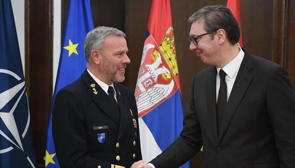 Poignée de main entre un officier de marine et un représentant civil, en présence des drapeaux de l'OTAN, de l'UE et de la Serbie.