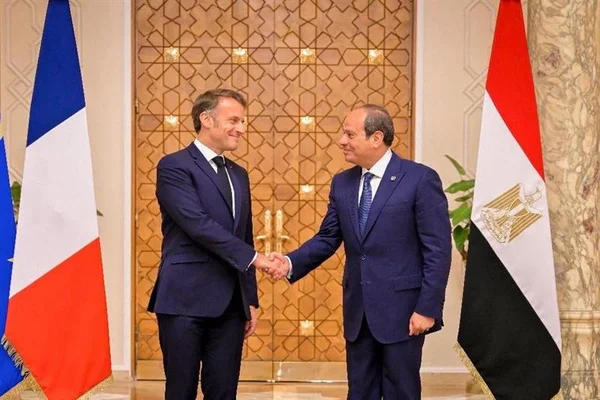 Emmanuel Macron et le président égyptien Abdel Fattah al-Sissi échangeant une poignée de main devant les drapeaux des deux pays.